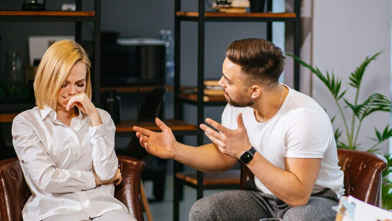 A man gestures with open hands while a woman sits beside him, looking down and crying.