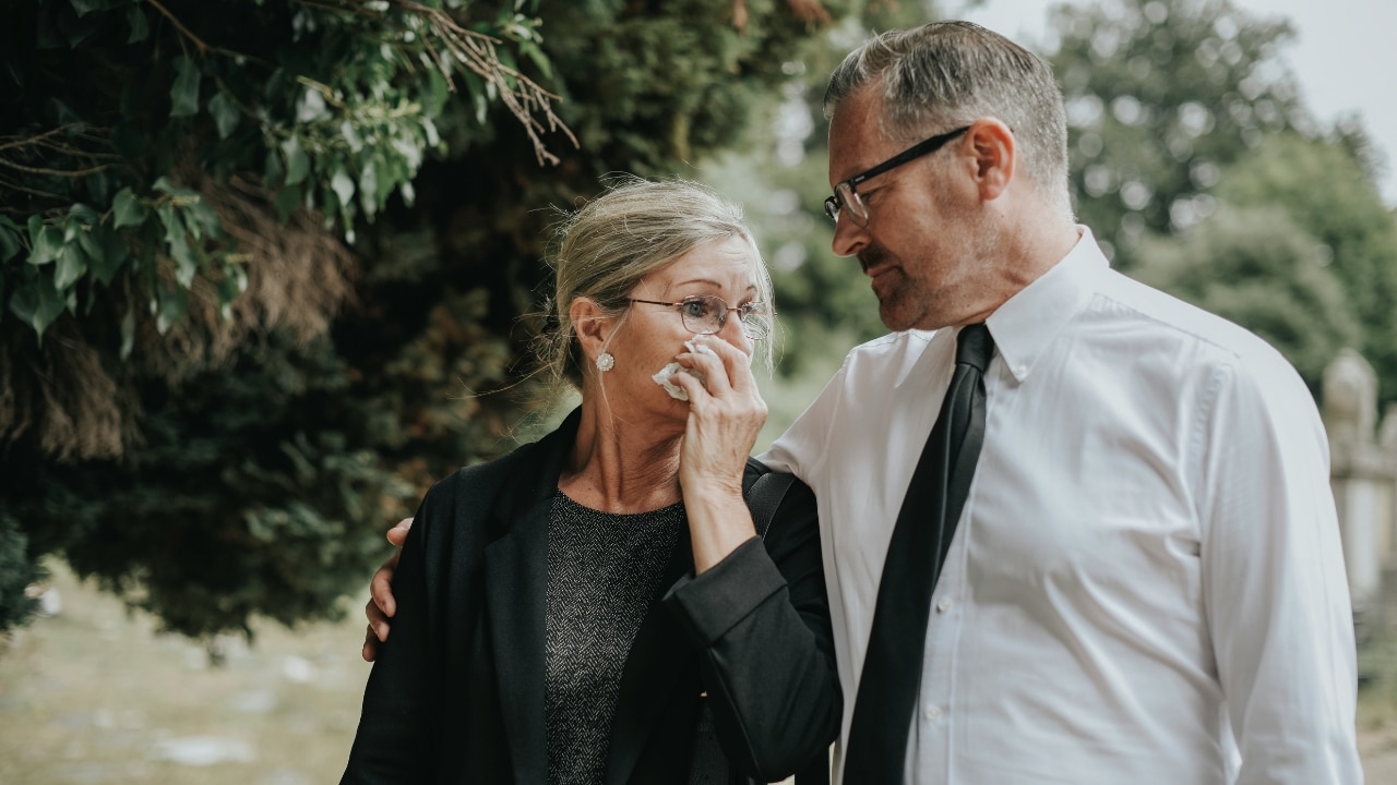 A man in a white shirt comforts a woman in black wiping her eyes outdoors.