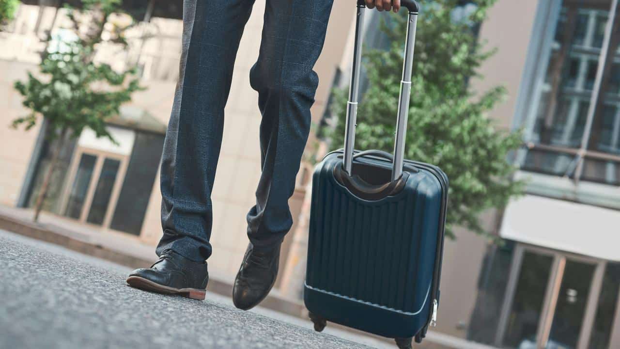 A man in dress shoes and trousers walking with a rolling suitcase.