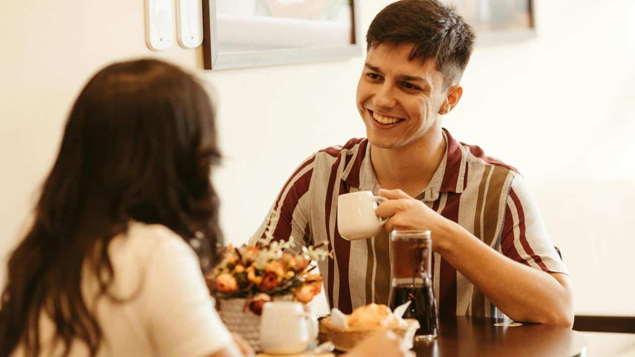A man smiling while having coffee with a woman at a café table.