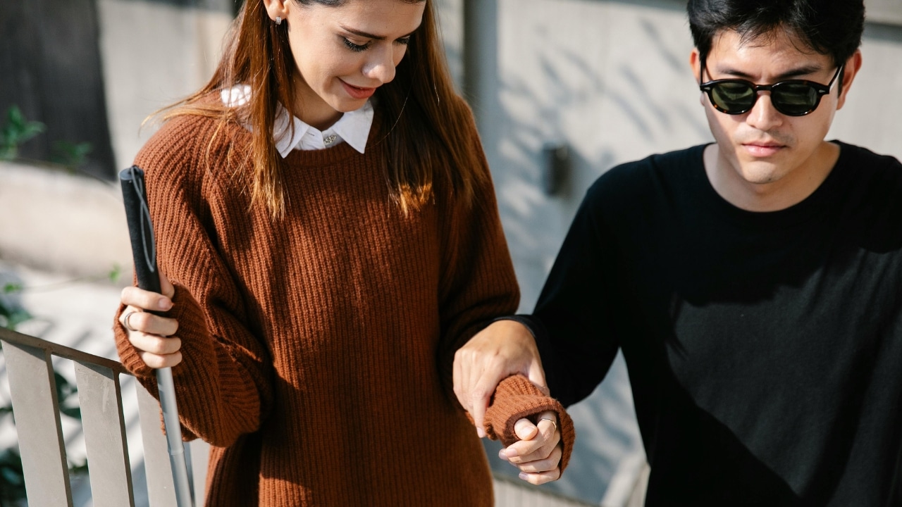A woman in a brown sweater holding a white cane while guiding another person’s arm.