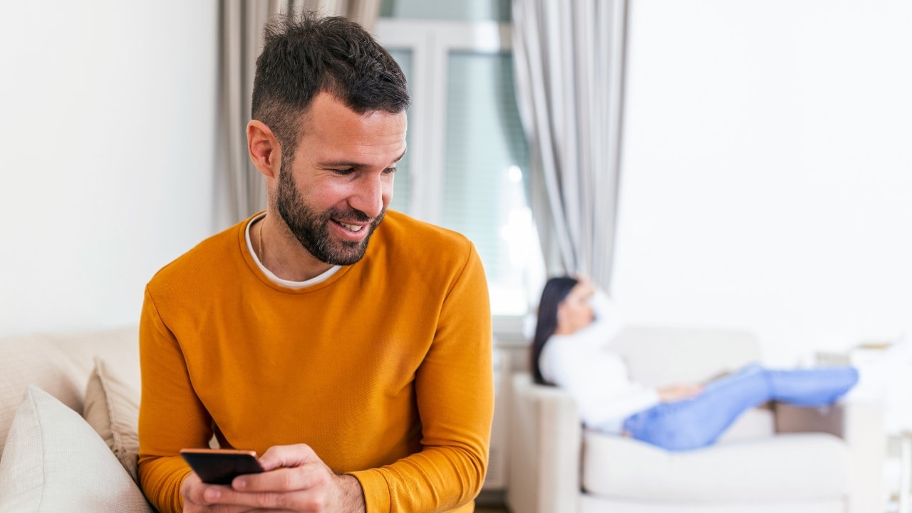 A man smiles at his phone while a woman sits blurred in the background sofa.