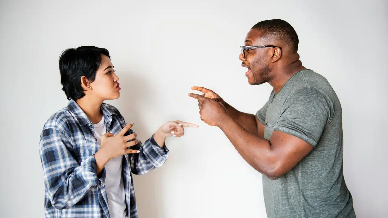 A woman in a flannel shirt and a man in glasses point toward each other.