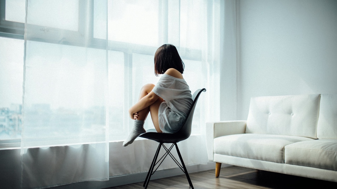 A young person with short, dark hair sitting alone on a chair, facing a large window covered by sheer curtains