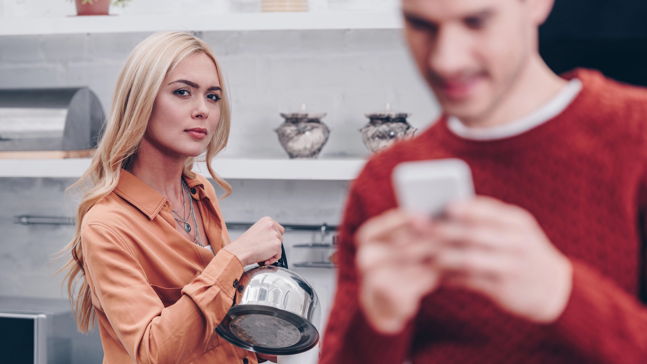 A woman holding a kettle and looking at the camera while a blurred man checks his phone.