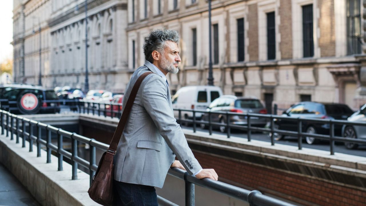 A middle-aged man in a gray jacket and shoulder bag looks pensively over a street railing.