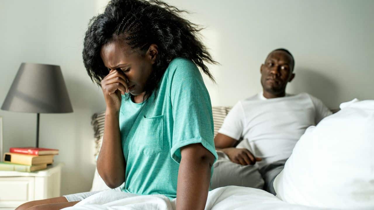 A woman sits on a bed covering her face while a man watches behind.