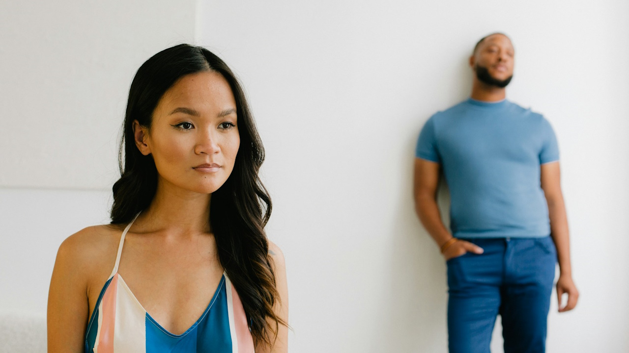 A woman with an inscrutable expression while a bearded man stands in the background.