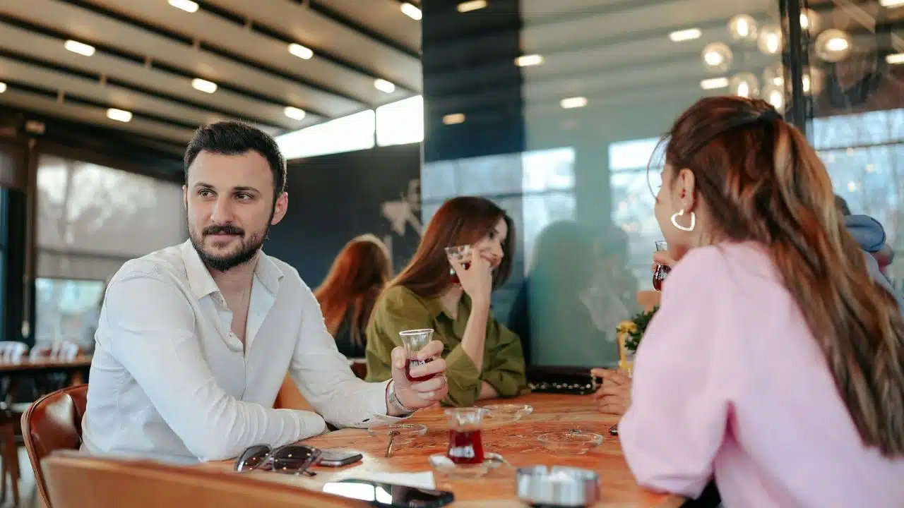 A bearded man sits at a table with three women, holding a small glass of tea.
