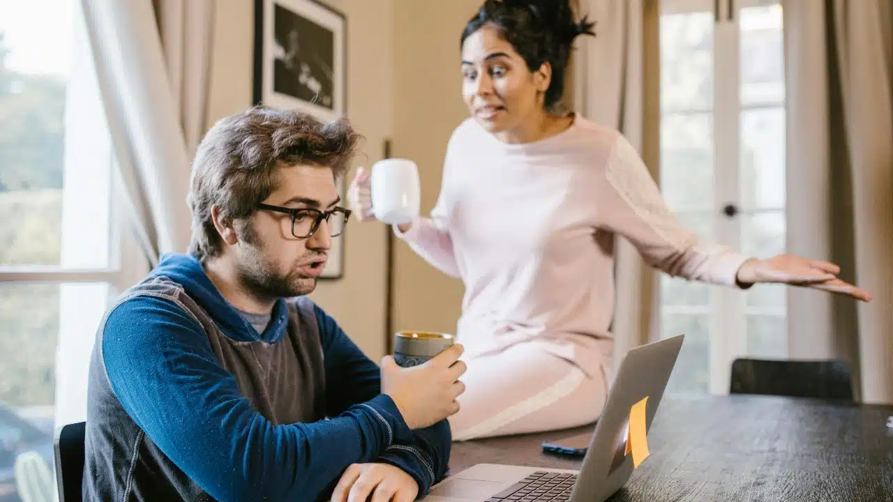 A man with glasses sits at a laptop while a woman stands behind him gesturing.