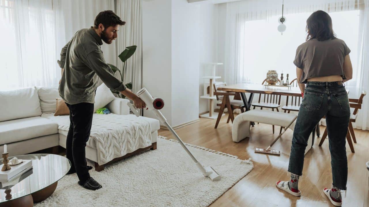 A man vacuums a white rug while a woman sweeps the floor in a bright living room.