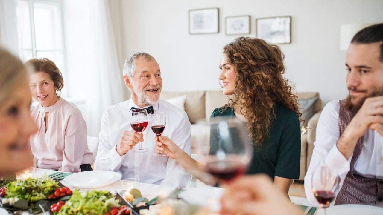 People sit at a dinner table smiling and clinking wine glasses together during a meal.