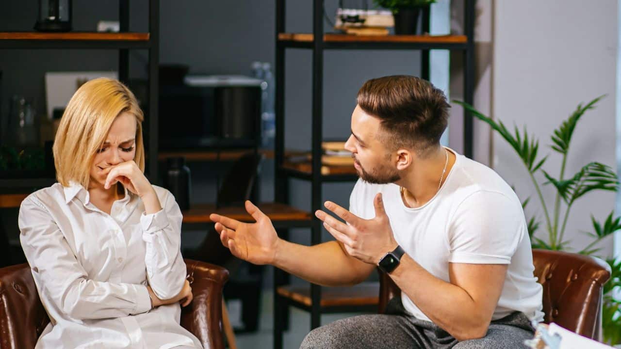 A man gestures with both hands toward a woman who is covering her mouth.