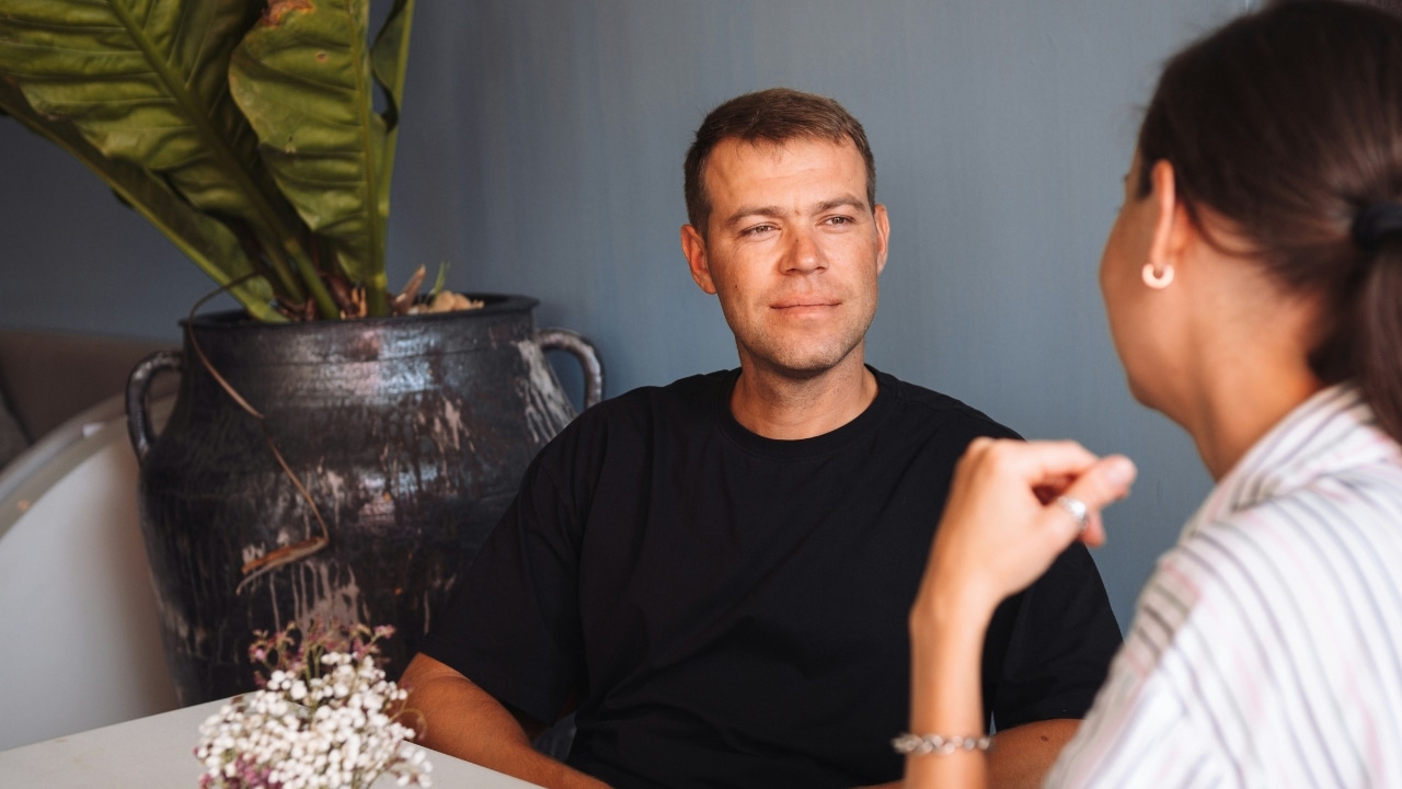 A man in a black shirt sits at a table, looking attentively at a woman.