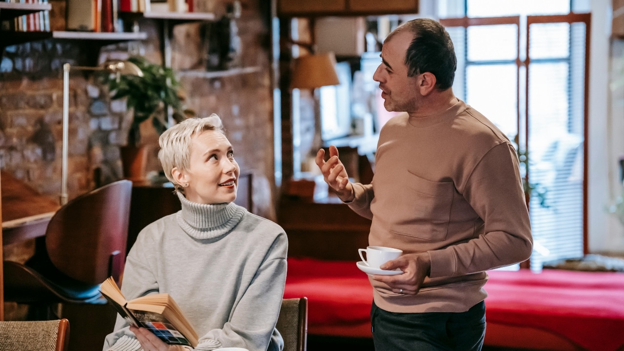 A man stands holding a teacup while talking to a seated woman holding a book.