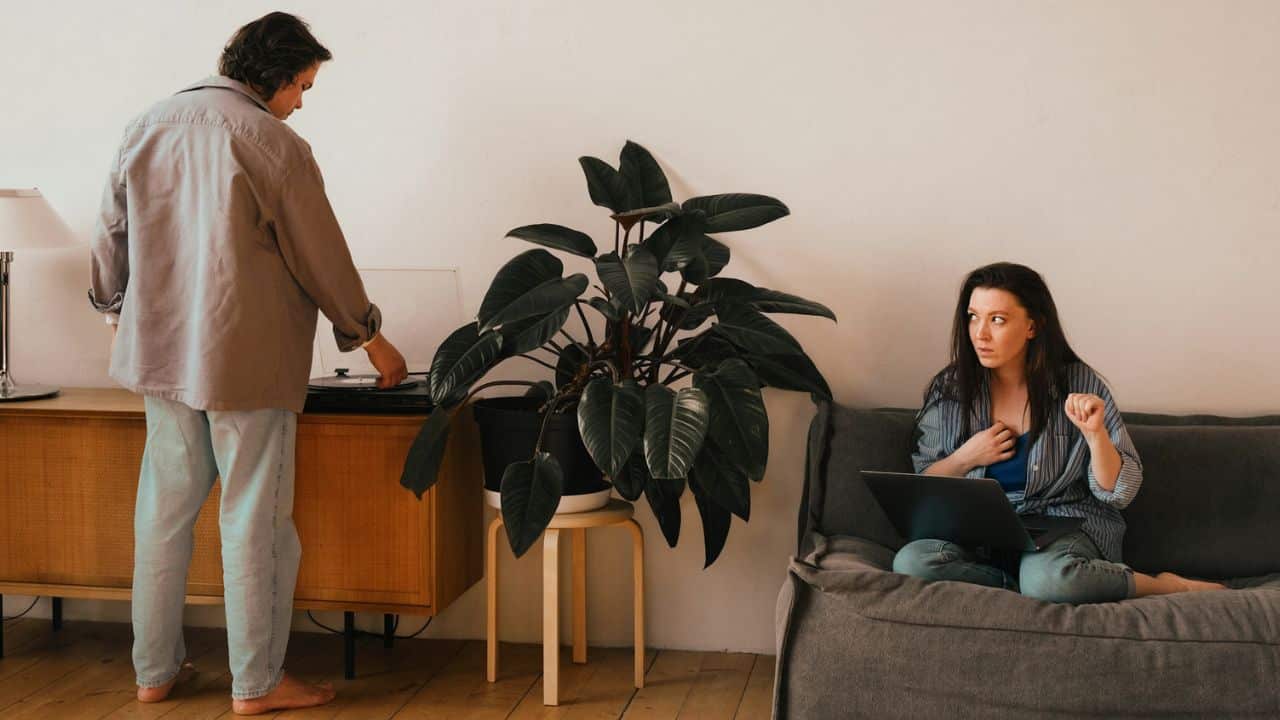 A woman sits on a sofa with a laptop while a man operates a record player.