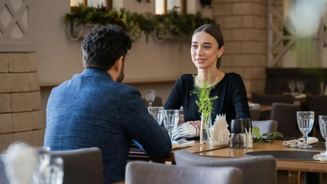 A woman in a black dress sits at a restaurant table looking across at a seated man.