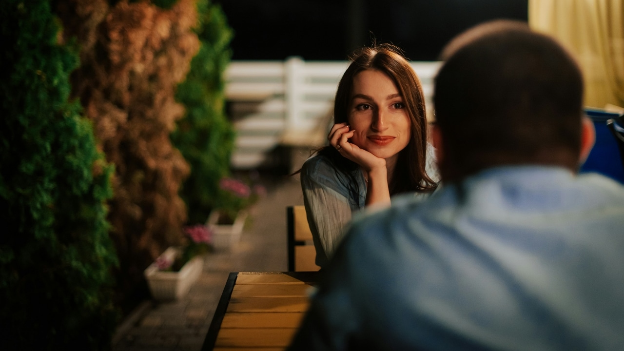 A woman rests her chin on her hand while looking at a man across a table.