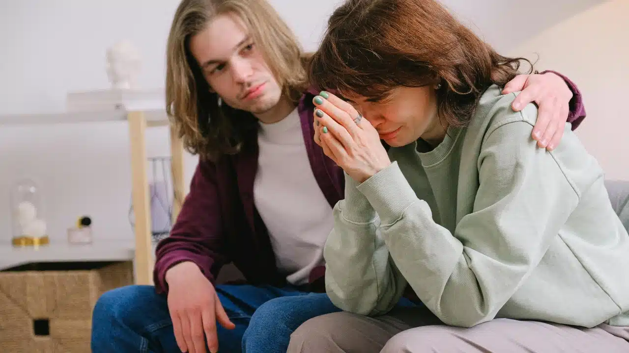 A person with long hair places a hand on the shoulder of someone who is crying.