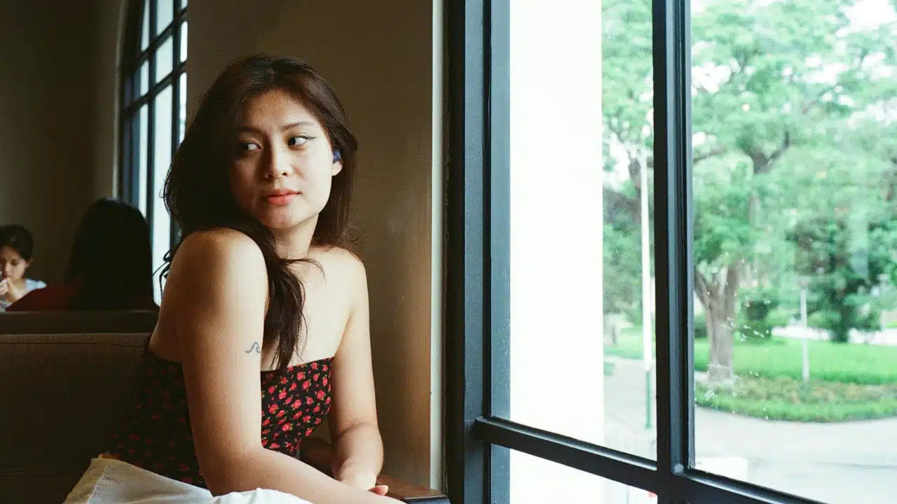 A woman in a floral top sits indoors and looks out of a large window.