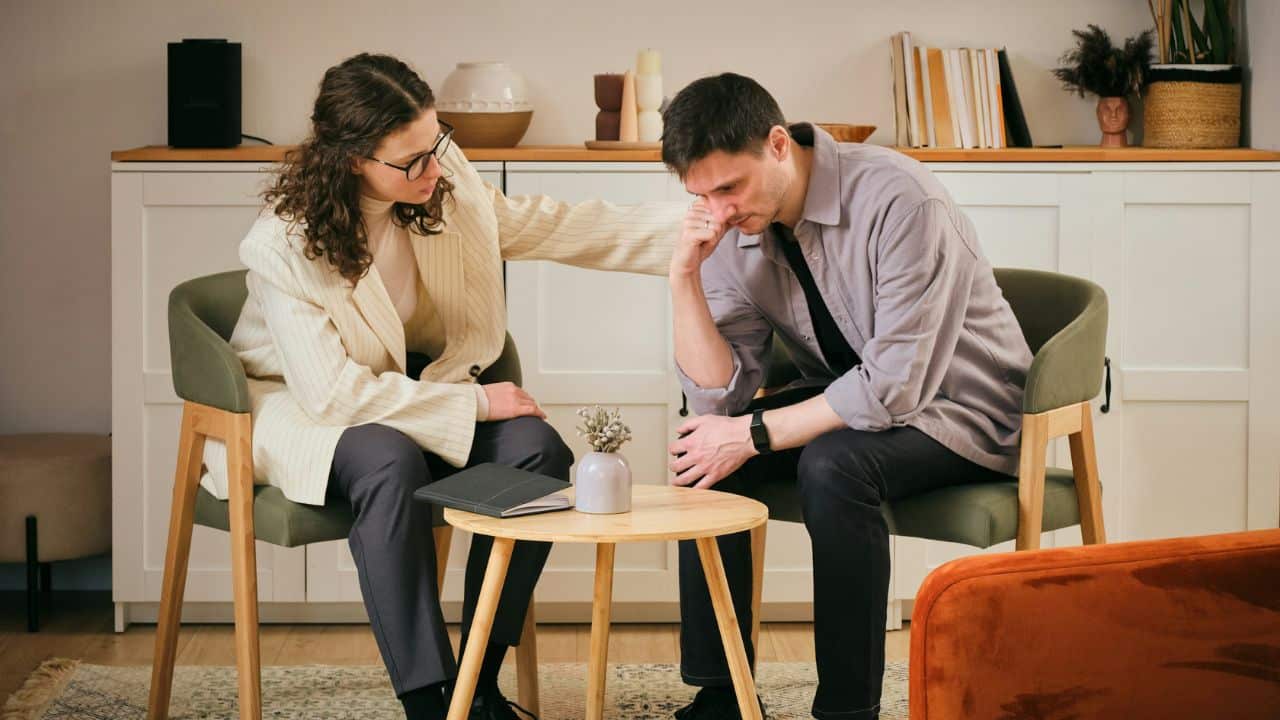 A woman in a white jacket places her hand on the shoulder of a seated man.