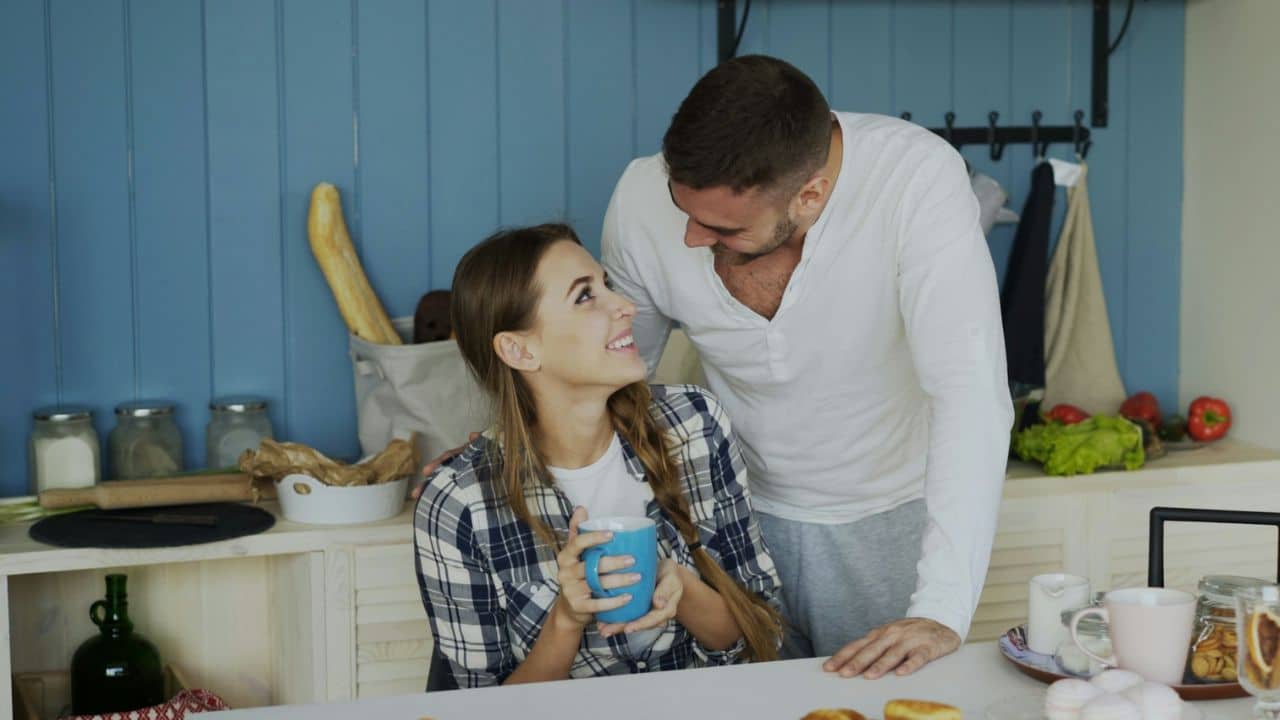 A man leans over a table to look at a smiling woman holding a blue mug.