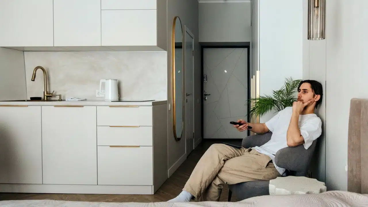 A man sits in an armchair holding a remote control in a modern studio apartment.