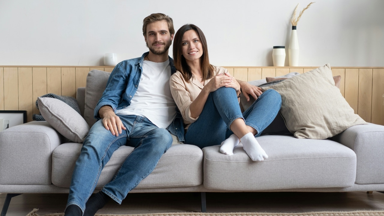 A smiling man and woman sit closely together on a grey sofa with several pillows.