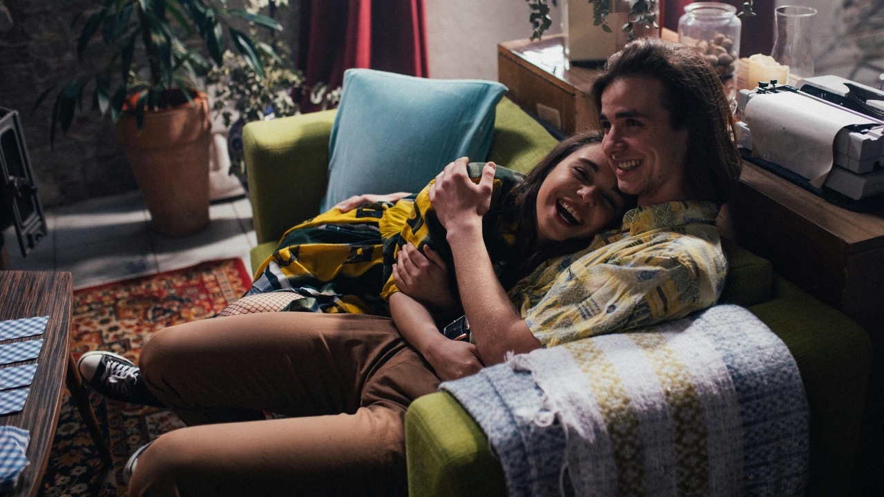 A man and woman sit closely together on a green armchair, laughing in a cozy room.