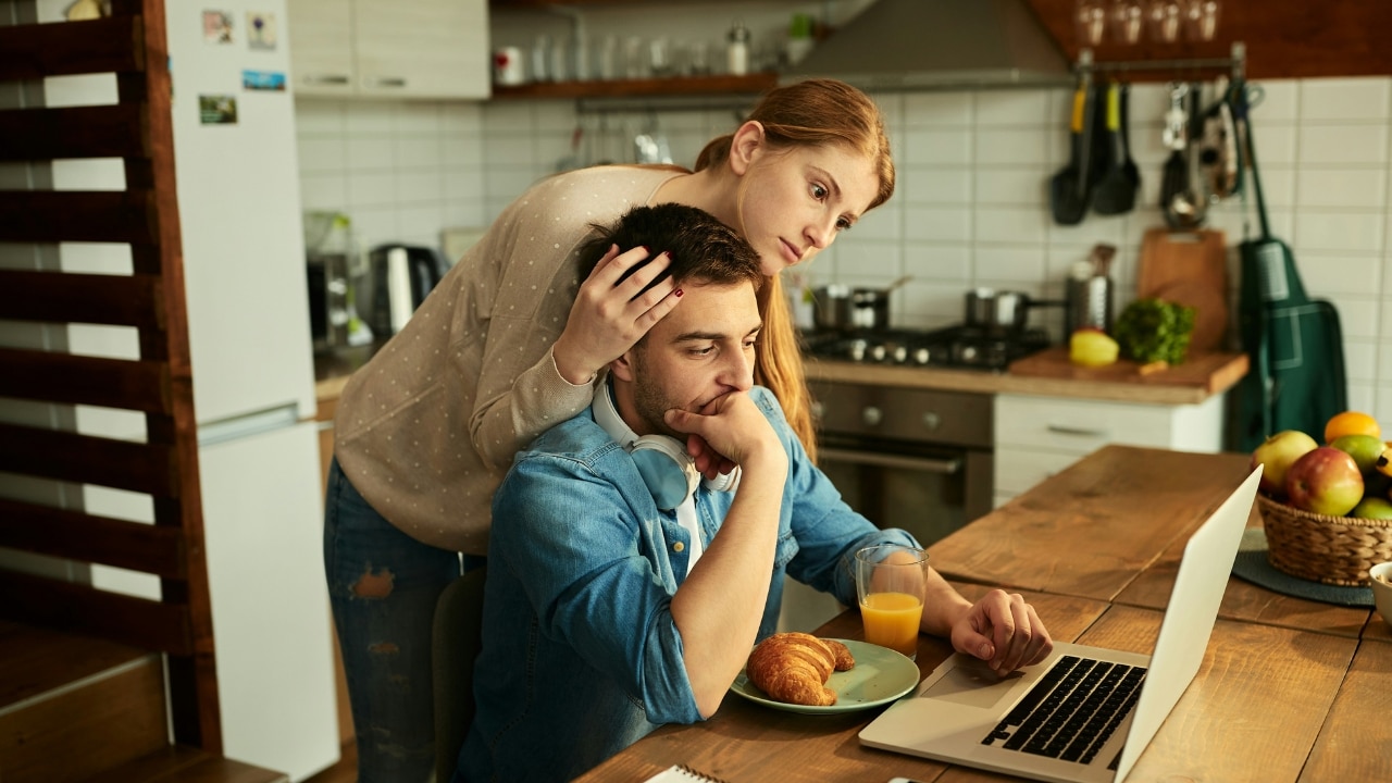 A woman leans over a man sitting at a laptop, placing a hand on his head.