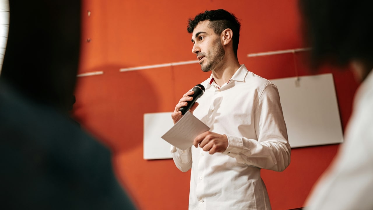 A man in a white shirt speaks into a microphone while holding a paper.