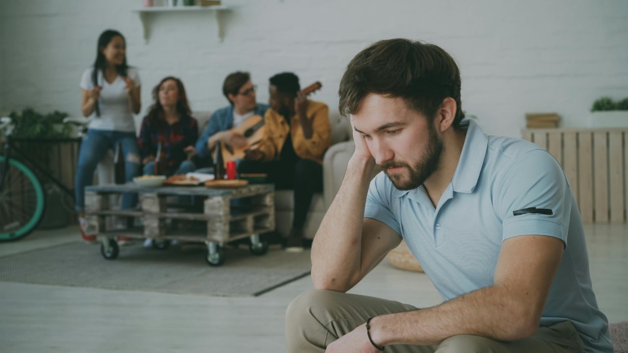 A bearded man sits in the foreground looking down while a group of people talk in the background.