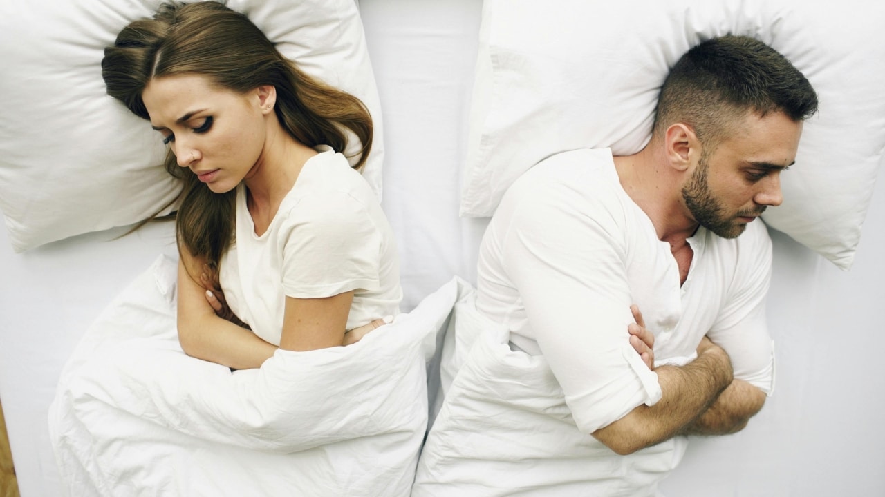 A man and woman lie in bed facing away from each other with arms crossed.